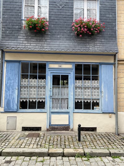 The image shows the facade of a building on a cobblestone street in Honfleur, France. The building has a grey slate roof and a cream-colored upper story with two windows, each adorned with a box of red and pink flowers. Below, the ground floor features a light blue door with a transom window and two large windows with light blue shutters. Both windows are dressed with white lace curtains. A small plaque above the door reads "16". To the left of the entrance is a grey utility access panel. To the right, a black bollard stands on the cobblestone street, next to a wrought iron grate set into the wall. The street itself is paved with unevenly sized cobblestones and large stone slabs. The overall scene evokes a charming, historic European streetscape.