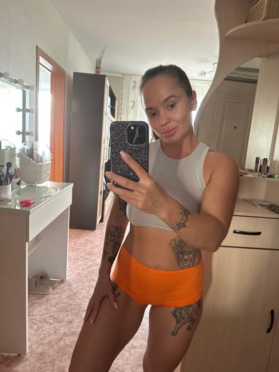 A woman with dark hair pulled back is taking a mirror selfie. She is wearing a white tank top and orange athletic shorts. She has tattoos on her arms and thighs, including a tiger on her right thigh and floral designs on her left thigh and arm. Her left hand is holding a black phone with a sparkly case. She is smiling at the camera. The background shows a dresser or closet with wooden doors and drawers, and a white vanity with makeup and brushes. The scene appears to be indoors, possibly a bedroom or dressing area, in a home. The lighting suggests it could be daytime. The location context is Yuzhnouralsk, Russia.