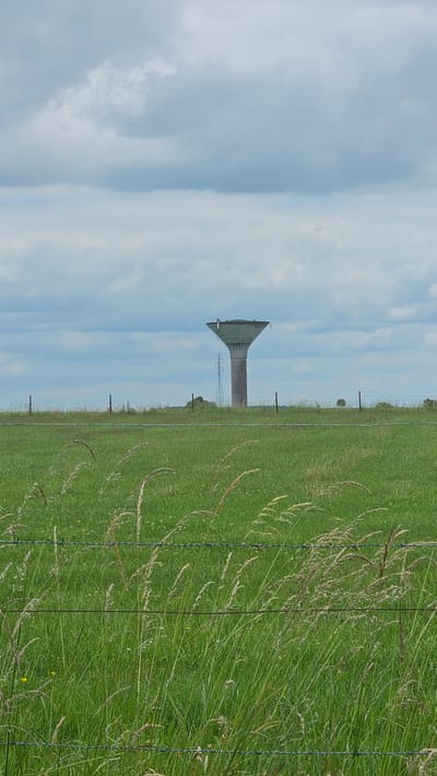 A water tower stands in the distance, centered against a cloudy sky. The tower is concrete, with a wide, flared top and a narrower, cylindrical base. It is located in a field of green grass, which is partially obscured by barbed wire fencing in the foreground. The field is dotted with small white flowers and taller stalks of dry grass. In the far distance, behind the water tower, a power pylon can be seen. The overall impression is one of a rural or semi-rural landscape under an overcast sky. The scene is located in Boussu-lez-Walcourt, Belgium.
