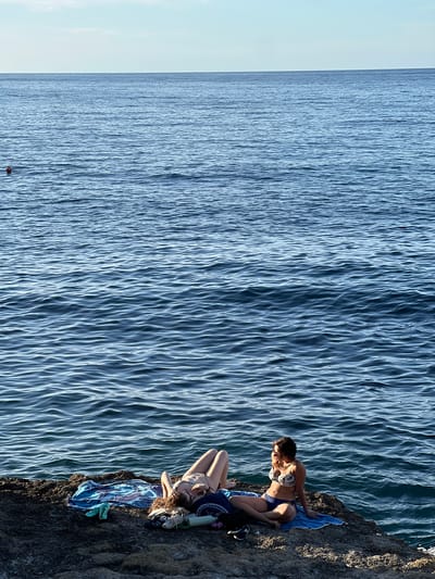 The image depicts a coastal scene in Sperone, Italy, featuring two individuals relaxing on a rocky shoreline adjacent to the sea under a clear sky. The horizon line is distinct, separating the light blue sky from the dark blue, rippled ocean. In the water, a small red buoy is visible on the left side, and a swimmer's head is partially submerged further out in the upper-left quadrant.
On the dark, rugged rocks in the foreground, two women are present. The woman on the left is lying on her back, facing upwards, on a multi-colored (blue, purple, white) towel. She is wearing a light-colored bikini and has curly reddish-brown hair. Beside her, a pair of light blue/green sandals, light-colored sneakers, and a dark piece of fabric or bag are placed. The woman on the right is seated upright on a blue towel, looking towards the right side of the frame, away from the camera. She is wearing a white floral-patterned bikini top and dark blue bikini bottoms, with sunglasses resting on her head. Near her feet are a dark bag and a pair of black sneakers. Both individuals appear to be sunbathing or relaxing by the sea.
