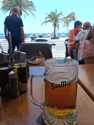The image presents a sunny, daytime scene from an outdoor cafe or restaurant in Puerto Del Carmen, Spain, with the ocean visible in the background.
In the foreground, a wooden table holds a clear glass mug of San Miguel beer, which has condensation on its surface, indicating it's cold. The mug clearly displays "CERVEZAS San Miguel SINCE 1890". A black coaster is beneath it. To the left, a metal caddy holds four condiment bottles, including one with red sauce, one with olive oil, and shakers likely for salt and pepper.
Beyond the table, a diverse group of people are seen. To the left, a man in a black polo shirt stands with his back to the viewer, looking towards the sea. To the right, several individuals are seated at other tables, including a woman in a pink top engaged in conversation with a bald man in a light-colored patterned shirt. Part of a child or another person in a floral-patterned top is also visible. Their activity suggests casual dining or socializing.
The background reveals a vibrant coastal setting. A street or promenade separates the cafe from the beach. Several tall, green palm trees are scattered throughout the scene. The sky is a bright, clear blue, meeting a tranquil azure ocean. On the beach, a few straw-thatched umbrellas are visible. A scooter is parked near the road. The overall atmosphere is relaxed and indicative of a holiday destination.