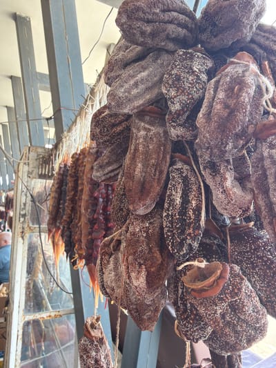 In Kareli, Georgia, dried persimmons and churchkhela are displayed in an outdoor market. The persimmons, a deep brown color with a powdery white coating, hang in clusters from strings. Adjacent to them, strings of churchkhela, resembling elongated sausages in various shades of red and brown, are also suspended. The background shows a metal structure, possibly part of a stall, with a person visible in the distance to the left. Sunlight illuminates the scene, highlighting the textures of the dried fruits.