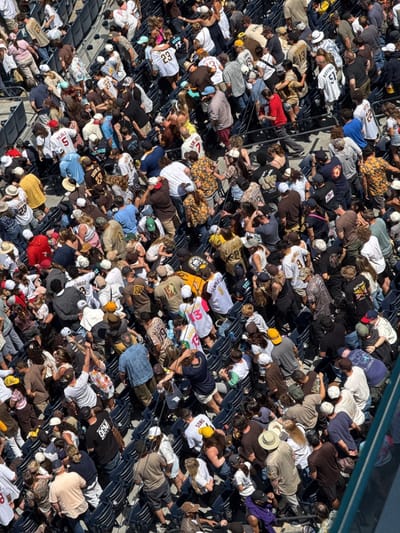 A high-angle, medium shot captures a crowded stadium setting, with numerous people occupying the blue seating. The scene is densely packed, with individuals sporting a variety of attire, including baseball jerseys, hats, and casual wear. The overall environment suggests a sunny day at a sports event, likely a baseball game given the prevalence of team apparel. The composition is busy, with movement and interactions amongst the attendees creating a dynamic atmosphere. The location is in San Diego, United States.