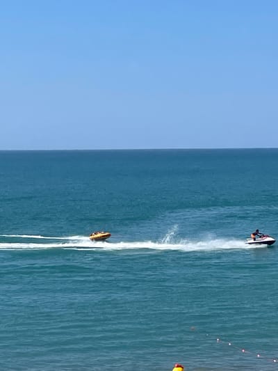 The image depicts a bright, sunny day at a beach in Giachrypsh, Abkhazia. The sea is a vibrant blue, with gentle waves. In the foreground, the edge of the beach is visible with some small buoys connected by a string. Two watercraft are active on the sea. On the right, a person is riding a jet ski, creating a white spray of water. To the left of the jet ski, an inflatable tube, also known as a "banana boat," is being towed, with several people riding on it. The sky is a clear, pale blue.