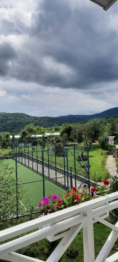 This is a vertical, eye-level shot of a fenced green sports court situated on a grassy lawn. The court is enclosed by a tall, dark green metal fence. A pathway made of flat, grey stones runs alongside the court, separated by a low stone wall. Beyond the court, the landscape is a vibrant green with trees and hills, some of which are covered in dense foliage. In the distance, a hazy mountain range is visible under a dramatic, overcast sky filled with dark, heavy clouds. In the foreground, a white railing with a planter box containing colorful flowers like pink, red, and purple geraniums, is present. The scene appears to be a recreational area or resort, possibly in a rural or mountainous setting. No people are visible. The setting is the outdoor area of a property in Ikindzhi Nyugedi, Azerbaijan.