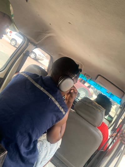 The image displays the interior of a worn vehicle, likely a commercial passenger van or bus, located in Jakande, Nigeria. The perspective is from the rear, looking towards the front.
In the mid-ground, a person with short dark hair is seated, wearing a dark blue sleeveless top with light-colored stripes across the shoulders and light-wash ripped denim shorts. This individual is wearing large, silver-grey over-ear headphones and has their right hand raised towards their ear or mouth.
Ahead of this person, the back of another passenger's seat is visible; its light-colored headrest is severely damaged with significant tearing and exposed foam. The passenger in this seat has dark, curly hair and is wearing a red shirt. A partial view of a third individual, wearing a green and beige cap, is visible in the upper left corner, possibly in the driver's or front passenger seat.
The vehicle's interior ceiling, or headliner, is light-colored, appears aged, and shows signs of sagging and minor imperfections. A blue-tinted visor or panel is visible above the front passenger side. Through the vehicle's windows, other vehicles are visible on a street or in a parking area, including a light-colored sedan to the left and a red vehicle in the distance through the front windshield.