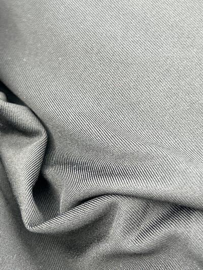 The image shows a close-up of a piece of gray fabric with a diagonal weave pattern. The fabric is draped and folded, creating shadows and highlights that add depth to the image. The weave pattern is visible throughout the fabric, and the texture appears soft and flexible. The image contains no information related to the location of Tlik, Armenia, or any other location, nor does it depict any people, subjects, objects, actions, interactions, or a specific environment.