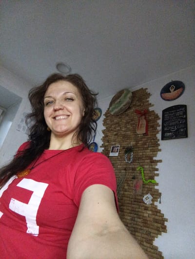 This is a selfie of a woman in Norilsk, Russia. The woman has long, dark, wavy hair and is smiling at the camera. She is wearing a red t-shirt with a white symbol on it. Around her neck, she has a pendant on a black cord. The background shows a wall decorated with wine corks arranged in a brick-like pattern. There are several items pinned to the cork wall, including a small framed picture, what appears to be a festive decoration, and a green, possibly beaded, object. Two circular wall decorations are also visible, one on the left depicting a dancer and another on the right with a dark background and an orange dancer. A black chalkboard-style sign with white writing is on the right side of the wall. The lighting suggests it is daytime indoors.