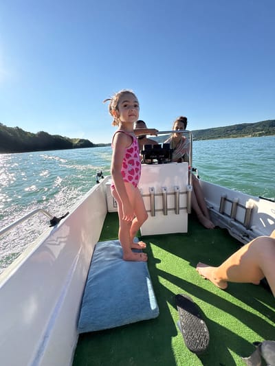 A low-angle, medium shot captures a young girl in a pink strawberry-patterned swimsuit standing on a boat's deck. She is facing forward and to the right, her gaze directed slightly upwards and away from the camera. Behind her, two adults are seated. A woman with dark hair pulled back in a bun is at the helm of the boat, wearing a light-colored, patterned garment. A man with dark skin and a shaved head is partially visible behind her. The boat is on a body of water, with the sun glinting off the surface. Green, hilly terrain lines the horizon under a clear blue sky. The boat's interior is covered with green carpeting, and several white rod holders are mounted on a console. A blue cushion is positioned on the deck near the girl. The image appears to be taken during daylight hours, with strong sunlight casting shadows. The location is identified as the city of Golyama Zhelyazna, Bulgaria.