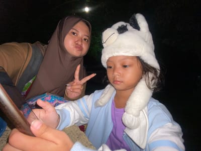 Two individuals pose for a photo at night in Kalitengah, Indonesia. The person on the left wears a white animal-themed hat with black spots, a light blue and white striped jacket over a purple shirt. The person on the right wears a brown hijab, displaying a peace sign. They are seated with a metal bar in the foreground. The setting is dark with sparse vegetation and distant lighting visible.