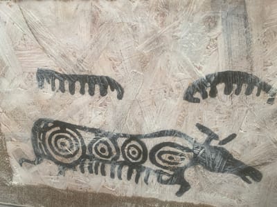 The image resembles cave paintings on a wood-grain surface. There is a large animal with concentric circles along its body, suggesting internal organs or spiritual symbols. Two smaller, indeterminate creatures are depicted above. All figures are drawn in black, contrasting with the lighter background. The style is crude and simple, mimicking ancient rock art.
