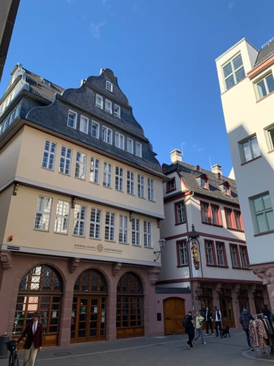 The image shows a street in Frankfurt am Main, Germany, at the intersection of Neue Mainzer Straße and Schillerstraße. The scene is an urban street in front of a building with an arched entrance and windows, with the words "Evangelisches Gemeindehaus" above the entrance. The street is paved and there are pedestrians walking on the sidewalk. To the right is a taller building, with windows and a white facade, that has a slight shadow cast upon it. There are trees in the distance, and a blue sky above. The scene is bright and sunny, likely taken on a clear day. There are several people in the street, most of whom are casually dressed. Two people are walking together on the left side of the street. Two people are walking together in the middle of the street, and a single person is walking on the right side of the street. Behind them is a group of people, about 6 in number, all walking together, likely a family or group of friends, and a dog walking with them. The building on the left has a large sign with a cross on it, suggesting it is a church or religious institution. The scene appears to be a snapshot of everyday life in Frankfurt am Main.