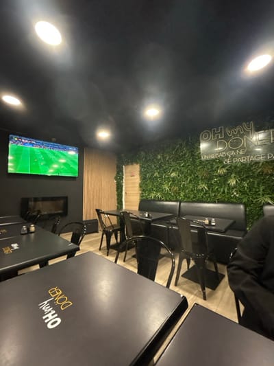 The scene depicts a modern restaurant interior. Black tables with branding are prominent, accompanied by black chairs. A long black banquette lines a wall adorned with artificial green foliage. A television displaying a soccer match is mounted on the opposite wall, which features a wood-paneled section. Overhead lighting fixtures illuminate the space.