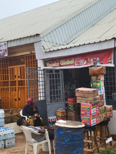 The image shows a storefront in Bokkos, Nigeria, with a red banner that reads "Emeka Global Ventures". In front of the store, there are stacks of water bottles and boxes of Indomie noodles. A woman wearing a pink headscarf is sitting in a white plastic chair, holding a baby. A young man is lying back in another white plastic chair, his eyes closed. The ground is dirt and gravel, and there are several metal barrels visible. The overall scene suggests a small business operating in an outdoor setting.