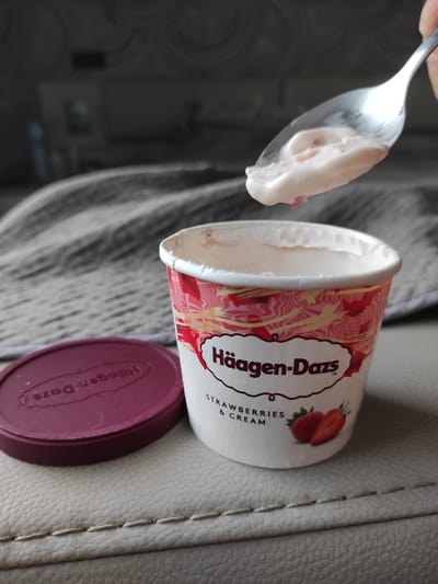 A close-up shot shows a spoon holding a scoop of pink ice cream over a partially eaten container of Häagen-Dazs Strawberries & Cream ice cream. The ice cream container is white with a red and pink swirling pattern. The lid, a deep red, is placed to the left of the container and has the Häagen-Dazs logo embossed on it. The scene appears to be indoors, possibly on a couch or upholstered surface, with a blurred background suggesting a domestic setting. The lighting is natural and diffuse, casting soft shadows.