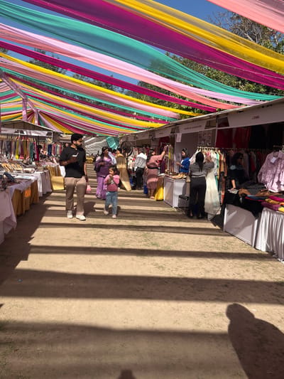 The scene is set in a marketplace in Sector 17, Chandigarh, India. The image captures a street lined with market stalls adorned with colorful fabric strips. The ground is a mix of dirt and gravel, with visible shadows cast by a canopy of colorful fabric strips.  There are several people walking along the street, some browsing the stalls. A young girl in a pink dress and a young boy in a black shirt are walking hand-in-hand on the left side of the image.  The market stalls are stocked with various products, including clothing, jewelry, and other items.  The air is clear and bright, with a blue sky and patches of cloud.  A large shadow can be seen in the center of the street.