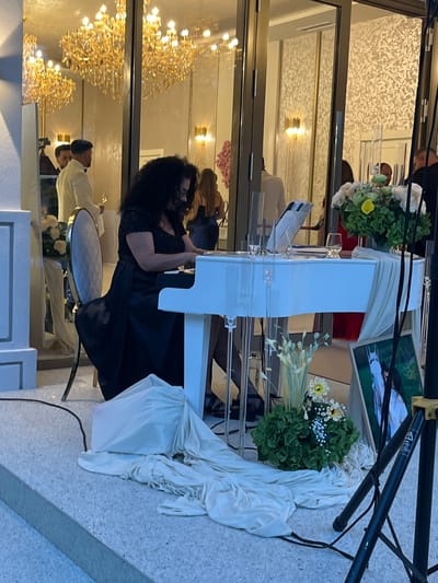 A woman with dark curly hair is playing a white grand piano in front of a large glass-enclosed building. She is wearing a black sequined top and a long black skirt. The piano has a clear stand and sheet music is visible on it. There is a floral arrangement next to the piano, with white and yellow flowers. A framed picture of a white horse is leaning against a speaker stand. The background shows chandeliers and people dressed in formal attire. The ground is tiled and there is a light-colored fabric draped over part of the piano and the floor. The scene appears to be an outdoor event or a building with large open doors.