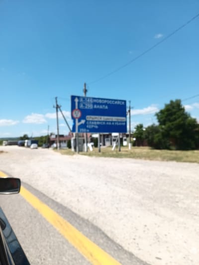 The image depicts a road intersection under a bright blue sky with scattered white clouds. A blue directional signpost stands prominently, indicating directions to various locations. Text on the sign includes "A-29 НОВОРОССИЙСК" (A-29 Novorossiysk) with an arrow pointing straight ahead, and "А-29 АНАПА" (A-29 Anapa) with an arrow pointing left. Below this, "КРЫМСК (центр города)" (Krymsk (city center)) is visible with an arrow pointing right, and further down, "СЛАВЯНСК-НА-КУБАНИ" (Slavyansk-on-Kuban) is also indicated with a rightward arrow. A circular red and white sign with the number "50" is attached to the signpost, likely indicating a speed limit.
In the background, several buildings with reddish roofs and some trees are visible, suggesting a rural or semi-urban setting. A few vehicles, including cars and trucks, are present on the road, suggesting ongoing traffic. The road surface appears to be a mix of gravel and asphalt, with a clear yellow lane marking visible in the foreground. The overall scene suggests a sunny day, possibly in the late morning or early afternoon, given the clear sky and bright lighting. The image appears to be taken from a moving vehicle, as evidenced by the perspective and the blurred foreground. The location context provided places this scene in Krymsk, Russia.