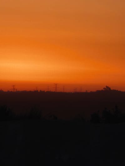 A photograph captures an orange sky at dusk over a silhouetted landscape. The sky transitions from a bright orange at the horizon to a softer orange towards the top of the frame. In the middle ground, the dark silhouette of a low horizon line is visible, punctuated by the outlines of several tall power pylons. Further to the right, the silhouette of a rocky outcrop with structures on top is also present. The foreground is entirely dark, obscuring any details of the ground or vegetation. The scene is likely an outdoor landscape, potentially in a semi-rural or industrial area given the presence of the power pylons. The location is Riba-roja de Túria, Spain.