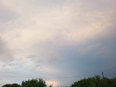 This is an outdoor, eye-level, wide shot of a cloudy sky with the tops of trees and a power line visible at the bottom of the frame. The sky is filled with soft, white, and pinkish clouds that transition to a pale blue on the right side of the image. A thin, dark power line stretches horizontally across the lower portion of the image. Below the power line, the dark green, leafy tops of trees are visible along the bottom edge. A wooden utility pole is visible on the right side, connected to the power line. The scene appears to be set in the city of Naruksovo, Russia.