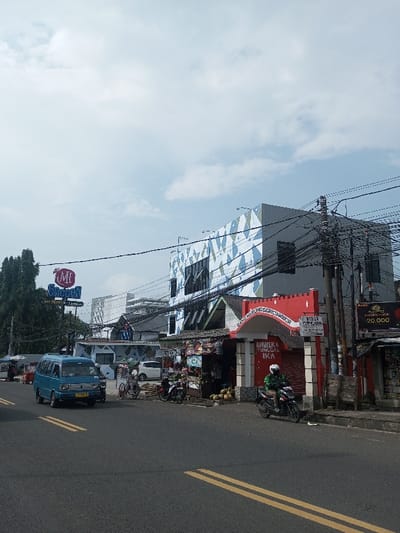 The image captures a bustling street scene in what appears to be an urban or semi-urban area during the daytime, under a partly cloudy sky. The perspective is from the road, looking towards a row of buildings and shops.
In the foreground, a paved asphalt road is visible, marked with two solid yellow lines down its center, indicating two-way traffic. The road appears dry and clear.
On the left side of the image, a light blue van, possibly a Suzuki Carry, is driving on the road. Behind it, a tall green tree line is visible. Above the trees and slightly to the left, a large sign with blue and white lettering reads "ME BACON" with smaller text below it that appears to say "AMT PEDAS". Further back on the left, other buildings can be vaguely seen.
Towards the center and right, a row of commercial buildings lines the street. A prominent multi-story building stands out with a distinctive geometric pattern of blue, white, and light green diamond shapes covering its facade. It has several dark, rectangular windows. In front of and below this patterned building is a lower structure with a red arched entrance and a red roof, resembling an entrance gate to a small market or alleyway. This structure houses several small shops or stalls. Piles of coconuts are visibly displayed for sale on the ground in front of one of these stalls.
A dense network of black utility poles and tangled electrical wires crisscrosses the mid-ground, connecting to the buildings and poles, partially obscuring the view of the architecture.
Several vehicles and people populate the scene. A small pushcart or street vendor's cart is visible between the blue van and the shops. On the right side of the road, a person wearing a green jacket and a white helmet is riding a black motorcycle, seemingly navigating past the shops. Another motorcycle is parked near the red-roofed structure.
Text visible in the image includes:
- On the large sign on the left: "ME BACON" and "AMT PEDAS".
- On the red arched entrance: "GANG WARUNG SERICIAMBOR" is clearly readable. Below the arch, a banner or sign displays "BHINEKA TUNGGAL IKA". To the right of the arch, a smaller sign reads "BIDAN" followed by blurred text.
- On a dark pole on the far right, an advertisement is attached, showing "BONU" (or similar) and a price, "Rp 20.000".
The overall impression is a lively street with ongoing commerce and daily activities, characteristic of a busy thoroughfare.