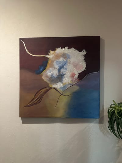 The image shows a square painting hanging on a textured, light beige wall. The painting depicts a stylized floral design with a cloud-like white and blue bloom on the left side against a maroon background. The right side features a gradient from blue at the top to brown at the bottom, with abstract lines and a brown leaf-like shape. A signature is visible in the upper right corner of the painting. A small potted plant with green, spiky leaves is positioned above and slightly to the right of the painting. The location is Jesús del Monte, Mexico.