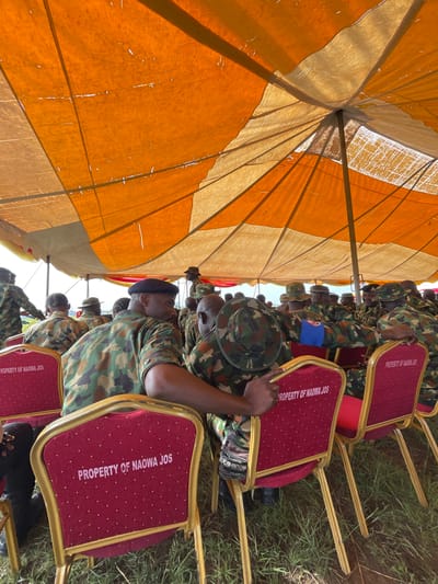 In Bahit, Nigeria, a group of individuals in camouflage military uniforms are seated on red chairs under an orange and tan tent. The chairs are labeled "PROPERTY OF NAOWA JOS." The people are facing away from the camera, with some interacting with each other. The environment appears to be outdoors, with grass visible around the chairs. The tent structure is visible overhead, supported by poles. The overall scene suggests an outdoor gathering or event.