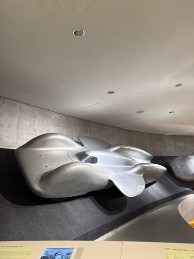 The image shows a museum exhibit of vintage Mercedes-Benz cars in Stuttgart, Germany. In the foreground, a silver, aerodynamic Mercedes-Benz W125 Rekordwagen, designed to break speed records, is displayed on a dark, textured platform. The car has a streamlined, teardrop shape with a visible cockpit and tail fins. To the right, partially visible, is another silver Mercedes-Benz vehicle, also with a rounded, aerodynamic form and the Mercedes-Benz logo prominently displayed on its front. The background features a textured concrete wall and a white, perforated ceiling with recessed lighting fixtures. A descriptive plaque is visible at the bottom of the image, indicating the year "1938" and "Mercedes-Benz Zw..." suggesting it is a Mercedes-Benz museum exhibit. The lighting is bright and even, typical of a museum setting, highlighting the metallic surfaces of the cars. There are no people visible in the frame.