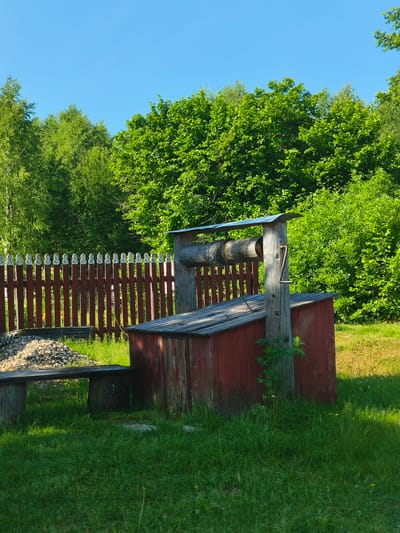 In the city of Zhukovo, Russia, a rustic wooden well stands in a grassy yard, bathed in the warm light of a sunny day. The well, constructed from weathered red planks and topped with a metal roof, features a wooden roller for drawing water. A metal crank protrudes from the wooden support beam. To the left of the well, a simple wooden bench sits beside a pile of grey gravel. Behind the well and bench, a red wooden fence with decorative white tops delineates the property. Lush green trees and bushes form a dense natural backdrop under a clear blue sky. The scene suggests a peaceful, rural setting.