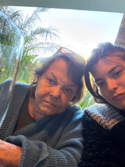 Two people are in a selfie taken from a low angle. The person in the foreground is an elderly woman with fair skin, wrinkles, and short brown hair, wearing glasses. She wears a grey sweater. Behind her, is a younger woman, with fair skin and dark hair wearing a patterned scarf with dark jacket. The background shows bright sunlight, palm trees, and a blurred glass object, partially obscuring the view, is likely a window. The location is presumed to be La Lucila, Argentina.