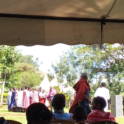The image shows a gathering in Keyian ward, Kenya. A group of people wearing traditional clothing are standing on a lawn. Several people, presumably audience members, are seated facing them. A man in a red shawl is walking towards a person in white. A white speaker is visible in the background. The scene appears to be outdoors under a tent or canopy. The environment includes green trees and grass.