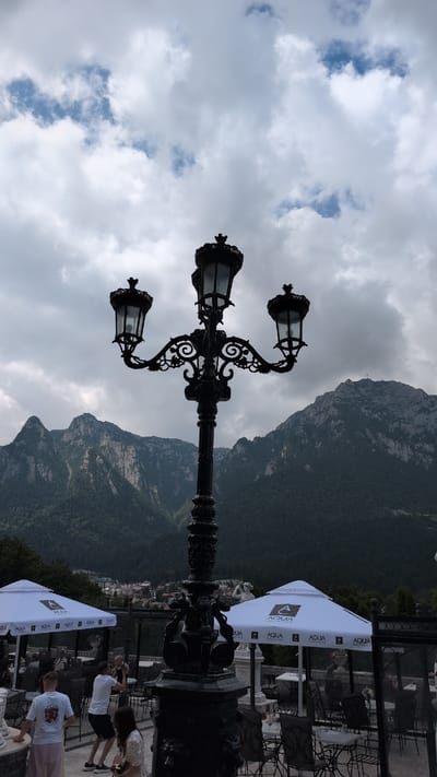 The image shows a view from a terrace in Bușteni, Romania, with a prominent black ornate lamppost in the foreground. In the background, a range of mountains covered in dense green forest rises, with rugged, rocky peaks visible in the upper portions. The sky is overcast with dramatic, heavy clouds.
On the terrace, several white umbrellas with the "AQUA CARPATICA" logo are set up over tables and chairs. Some people are visible, including two men in white t-shirts and shorts standing near the left side of the frame. One man is holding a baby. Another woman is sitting on the right side of the frame. The setting appears to be an outdoor dining area or a viewpoint cafe. The architecture of the terrace and some decorative statues are also partially visible.