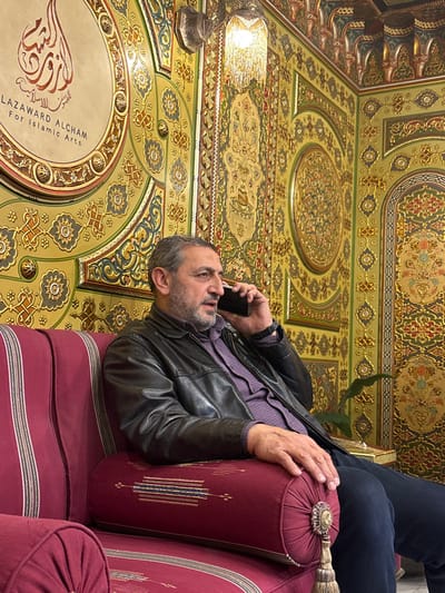 A man in a leather jacket and blue pants sits on a maroon striped couch, holding a phone to his ear. He has short gray hair and a beard. He leans against a maroon bolster pillow with tasseled ends. Ornate geometric designs in gold, brown, and green cover the ceiling and surrounding surfaces. A light fixture hangs from the ceiling. A round plaque is visible, containing Arabic text and the phrase "LAZAWARD ALCHAM for Islamic Arts".
