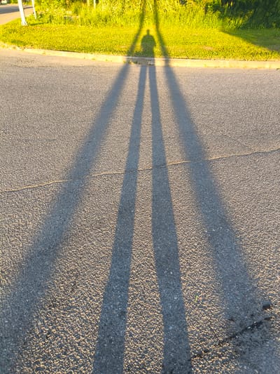 The image shows long shadows cast on an asphalt road, stretching from a grassy area with sunlight. The shadows suggest the presence of at least one person, possibly two, standing near the edge of the grass and curb. The sunlight illuminates the grass and the top of a curb, creating a stark contrast with the shaded road. The asphalt surface shows some wear, with a visible crack running across it. There are no people or identifiable structures visible on the road. The overall scene is one of an outdoor, daytime setting, likely in a public area with a road and adjacent green space.