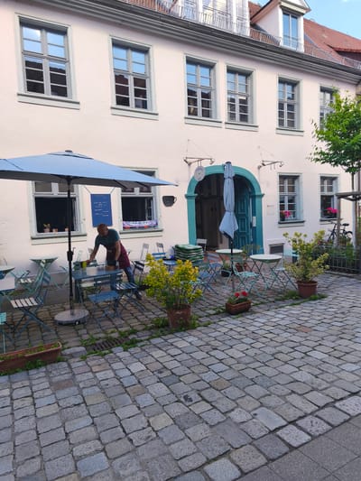The image shows the outdoor seating area of a cafe, likely located in Erlangen, Germany, based on the cafe's signage which reads "Steffis Cafe". A man, wearing a green shirt and a maroon apron, is seen tending to a table. He appears to be setting up or cleaning. Several small, round tables with blue metal chairs are arranged on a cobblestone patio. A large, closed blue umbrella stands centrally in the seating area. Potted plants with yellow and red flowers add a touch of greenery. The building facade is light pink with several windows. An arched doorway with teal trim leads into the cafe. A bicycle is parked to the right of the doorway, next to a fence. The scene suggests a typical European cafe setting.