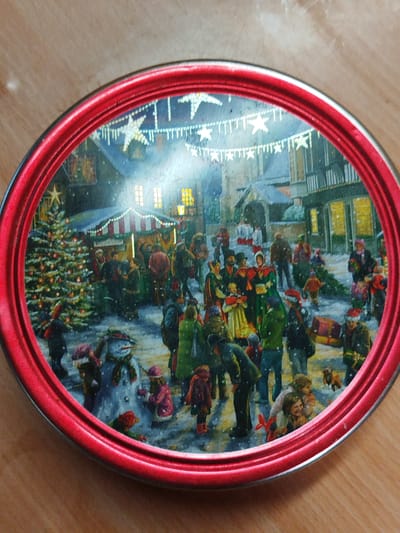 This image shows a painting of a Christmas market scene, likely on the lid of a tin. The setting appears to be a European town during winter, with snow falling. Several people are present, engaging in various activities. There is a decorated Christmas tree, a market stall with red and white striped awning, and buildings decorated with lights. Some people are buying goods, while others are caroling or carrying Christmas trees. There is a snowman in the foreground. Decorations including star-shaped lights strung across the square create a festive atmosphere. The location is NOT Боровково, Russia.