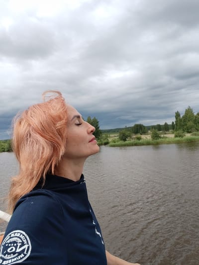 A side profile of a woman with peach-colored hair is captured as she looks upwards with closed eyes against a backdrop of a gray, cloudy sky. She is wearing a dark blue shirt with white lettering on the sleeve. The background features a calm body of water, with trees and greenery on the opposite shore, suggesting a natural, possibly rural, setting near Vytegra, Russia. The water's surface is disturbed by small ripples, and the sky is overcast, indicating a cloudy day.