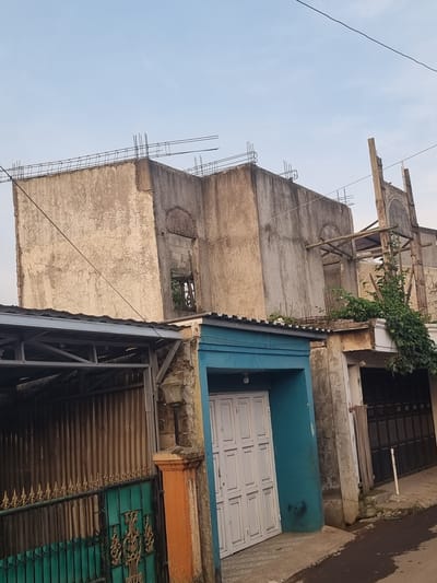 The image shows a multi-story building under construction in what appears to be a densely populated area of Cibodas, Indonesia. The building is made of concrete and has exposed rebar on the upper floors, indicating ongoing construction. The exterior walls are unfinished and show signs of weathering. Next to the building is another structure with a white door in a blue frame. Below is a metal roof with a partially visible green gate. The sky is visible in the background. An electrical wire runs across the top left of the image.