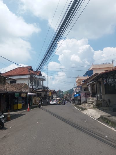The image depicts a street scene in what appears to be a Southeast Asian town, characterized by dense overhead power lines, narrow roads, and a mix of residential and commercial buildings. The road is paved with asphalt and shows signs of wear, with visible cracks and patches. On either side of the road are buildings of varying styles and conditions, from simple structures to multi-story buildings. Some buildings have tiled roofs, while others have more modern facades. Small shops and businesses line the street, indicated by signage and storefront displays. Vehicles, including motorcycles and cars, are present on the road. In the background, there is a hint of a hillside. The sky is partially obscured by clouds, suggesting a partly sunny day. The OCR text identifies some of the shop names and details, such as "Paradise," "BIDAN," and "GOONER Barbershop."