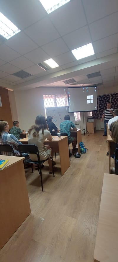 A group of people are in a classroom setting. In the foreground, a person with long brown hair wearing a light-colored dress is sitting at a desk. To their left, a person wearing a plaid shirt is seated. In the middle ground, several other individuals are seated at desks. A projector screen is at the front of the room, displaying a graphic with four squares. A man in a striped shirt is standing next to the projector, facing the seated individuals. The room has wooden flooring and a drop ceiling with fluorescent lights. The overall scene depicts a lecture or presentation. This scene is located in Tetiiv, Ukraine.