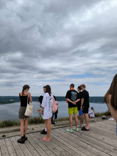 The image depicts a scenic overlook in the Horaivka area of Ukraine, providing a panoramic view over the Dniester River. The sky is largely overcast with varying shades of grey clouds, though hints of blue are visible towards the distant horizon above the river. The wide Dniester River flows through the midground, its banks lined with dense green vegetation.
In the foreground, a light-colored wooden deck or platform serves as the viewing area. Nine individuals are present on or near this platform. On the wooden deck, a group of six stands, primarily facing towards the river. This group includes two females: one in a black tank top, an olive-green skirt, and carrying a light tote bag; the other in an oversized white t-shirt with black graphics, pink flip-flops, and a pink backpack. Among the males in this group, one wears bright green shorts and light green clogs, another a light brown cap. To the immediate left and far right, two additional individuals are partially visible. Further back and to the right, on a grassy slope, one person sits on the ground facing away from the camera. The individuals appear to be observing the natural landscape.