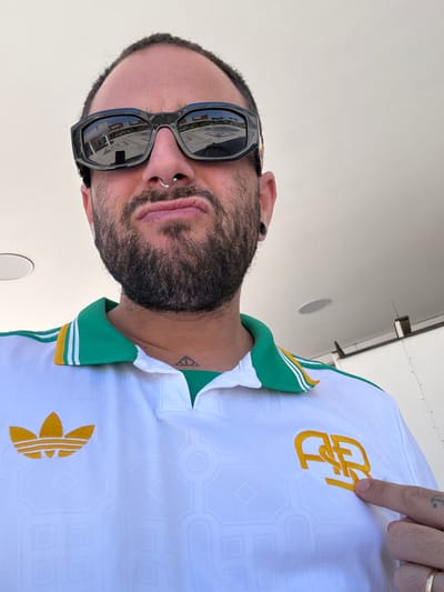The photograph shows a man with a beard, wearing sunglasses and a white shirt with green and gold trim. The shirt features an Adidas logo and another logo with intertwined letters. He is pointing at the logo with his right hand, which has a tattoo of an anchor on the wrist and a gold ring on the finger. The man also has piercings in his nose and ear, and a small tattoo on his neck. The background appears to be a plane interior. The photograph was taken in Pomezia, Italy.