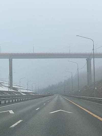 The image depicts a highway in Сапуголи, Russia, under foggy conditions. The road has two lanes with white directional arrows and a yellow line marking the right edge. A large bridge structure spans overhead, supported by tall concrete pillars. Streetlights line both sides of the road. The roadside slopes are covered with sparse vegetation and patches of snow. Visibility is significantly reduced due to the heavy fog.