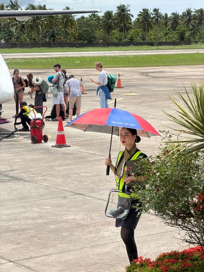 The scene appears to be at the Del Carmen Airport in the Philippines. A line of passengers disembarks an aircraft on the tarmac. An airport employee wearing a high-visibility vest holds a blue and red umbrella marked "FOR RAMP USE ONLY" and a clear bag. There are traffic cones, luggage, and a small cart near the aircraft. Tropical vegetation surrounds the tarmac area.