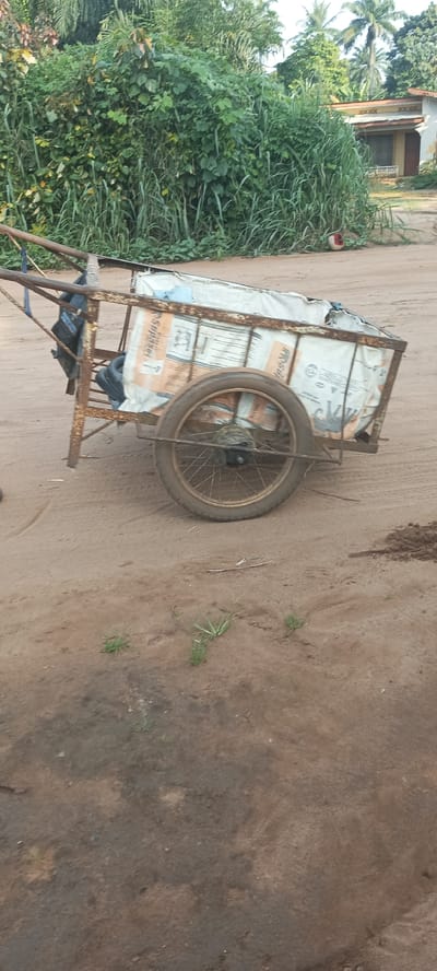 The image depicts a rusty, metal cart with one visible wheel, partially filled with bags. The cart appears to be parked on a dirt road. In the background, there is lush green vegetation and a glimpse of a house. The location is Amauzu, Nigeria. There are no people immediately visible.