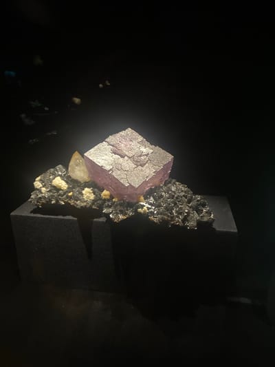 The image shows a mineral specimen, likely fluorite, displayed in a dark environment. The main feature is a large, cubic crystal, possibly purple or pink in color with a silvery sheen on the surface. This crystal is situated on a matrix of smaller, darker, crystalline material. The specimen appears to be mounted on a black, geometric support. The background is mostly black, suggesting a display case or dark room. Based on the prompt, this image may have been taken in Grenoble, France.