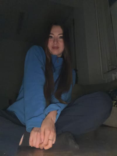 The image shows a young woman in a dimly lit room, possibly a living room or bedroom, based on the presence of a couch and what appears to be a radiator or air conditioning unit on the wall in the background. The woman has long, dark brown hair and is wearing a blue sweatshirt. Her hands are clasped together in front of her, and she is wearing a ring on her right hand. She is looking towards the camera with a slight smile. The overall lighting is low, suggesting it might be nighttime or a dimly lit interior. There are no visible signs of weather conditions or specific time of day beyond the lighting. The setting is domestic. There is no visible text in the image.