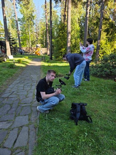 The image shows an outdoor scene in what appears to be a park or recreation area in Repino, Russia. Three people are present. One man is sitting on a stone-paved path holding what looks like a camera rig. Another individual is bending over, also appearing to be working with a camera or filming equipment on the grassy area adjacent to the path. A third person, wearing a tie-dye hoodie, is standing further back on the grass. A black backpack sits on the grass near the individual bending over. The area includes trees and a playground structure is visible in the background. The lighting suggests it is daytime.