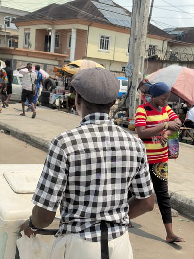 The scene depicts a bustling street in Jakande, Nigeria, on a clear day. In the immediate foreground, a dark-skinned male is viewed from behind, wearing a grey herringbone flat cap, a short-sleeved black and white plaid shirt, and light-colored trousers with a black strap hanging from the waist. His left wrist bears a black watch, and he grips a light-colored, rectangular object, possibly a cooler or vendor's cart, at the bottom left.
To the right of the foreground subject, a dark-skinned female, seen from the waist up, wears a dark blue headscarf, a red and yellow striped short-sleeved shirt, and black pants. Gold-colored bangles adorn her left wrist as she holds a colorful patterned plastic bag in her left hand, her right hand near her left forearm. She wears black sandals.
The midground features a concrete curb separating the subjects from a street populated by pedestrians and roadside activity. A tall, grey utility pole stands centrally, covered in wires and bearing a faded sign. Behind the female, a large, light-colored umbrella with red/orange stripes provides shade. Further down the street, multiple individuals are visible, including a man in a white t-shirt scratching his head, and a light-colored sedan parked on the left. Several makeshift stalls display goods, including stacks of tires and various merchandise under a yellow awning.
The background is dominated by a two-story, pale yellow building with a dark, sloped roof fitted with multiple solar panels. White columns and numerous windows are visible on the building's facade. Another building is partially visible to the right. The sky appears bright and somewhat hazy.