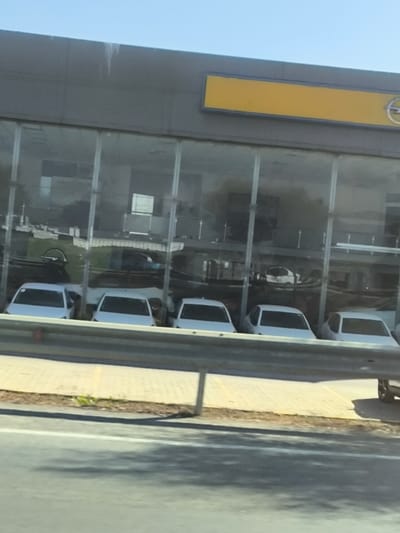 The image depicts an exterior view of a car dealership in Gonyeli, Northern Cyprus / Cyprus, observed from an adjacent roadway. The dealership building features a large, multi-pane glass facade on its lower level, behind which several new, white sedan vehicles are parked in a row. Reflections on the glass obscure some interior details but reveal additional vehicles and internal structures. Above the glass section, the building has a grey, solid upper facade, featuring a prominent rectangular yellow sign on the left and a circular Opel logo with its distinctive "Z" shape on the right.

In the foreground, a two-bar grey metal guardrail separates the viewer's position from the dealership's premises. Beyond the guardrail, a light-colored paved sidewalk, constructed from rectangular pavers, runs parallel to the building. Sparse green vegetation is visible between the sidewalk and the darker asphalt road surface in the immediate foreground. The sky above the building is clear and blue, indicating daytime conditions. No people are visible in the scene, nor are any specific actions or interactions depicted.